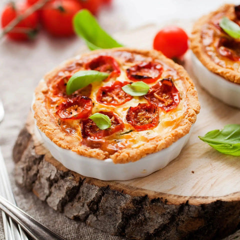 Small savory pies topped with cherry tomato slices and basil leaves, placed on a wooden platter.