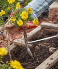 Using Premium Stainless Steel ComfortGEL All In One Hoe Cultivator With Comfy Grip For Weeding in garden bed with yellow flowers.