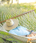 Person relaxing in a 13 Ft Double Cotton Rope Hammock For 2 450 Lb Capacity Free Extension Chains And Tree Hooks Hand Woven Traditional Style Designed In The USA Ultra Soft 8 mm Rope For Cozy Outdoor Lounging And Chillin, wearing a straw hat in a sunny garden.