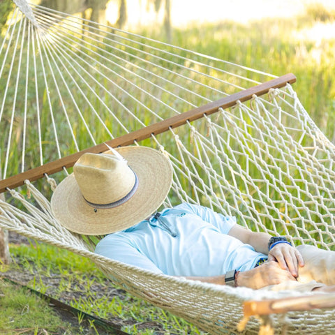Person relaxing in a 13 Ft Double Cotton Rope Hammock For 2 450 Lb Capacity Free Extension Chains And Tree Hooks Hand Woven Traditional Style Designed In The USA Ultra Soft 8 mm Rope For Cozy Outdoor Lounging And Chillin, wearing a straw hat in a sunny garden.