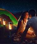 12 Pack Solar Torch Lights With Flickering Flames for outdoor use beside a couple camping near a tent in dim light setting.