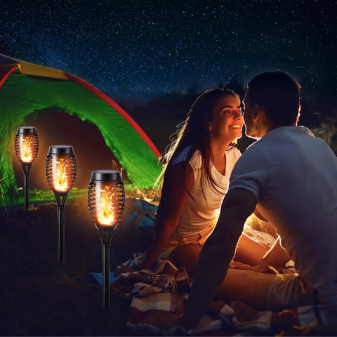 12 Pack Solar Torch Lights With Flickering Flames for outdoor use beside a couple camping near a tent in dim light setting.