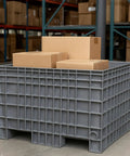 Dang Tough Heavy Duty Plastic Bulk Storage Tote, 42X29X28 Inch Stackable Pallet Box in gray with cardboard boxes on top.