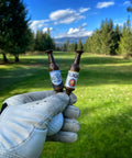 Gloved hand holding two Beer Bottle Golf Tees Pack Of 6 For Golfers - Virtually Unbreakable Recyclable Plastic - 3-1/4 Inch Tall on golf course.