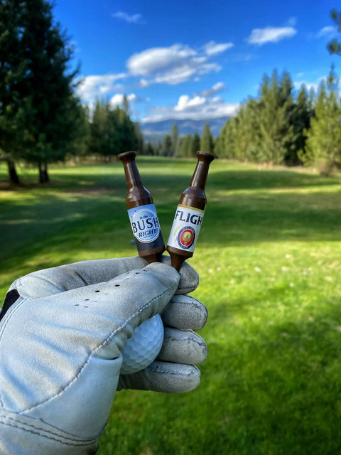 Gloved hand holding two Beer Bottle Golf Tees Pack Of 6 For Golfers - Virtually Unbreakable Recyclable Plastic - 3-1/4 Inch Tall on golf course.
