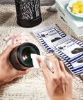 Lens cleaning tissue sheets and double-sided cloth kit; hands wipe a camera lens with tissue, 2 blue-striped booklets nearby.