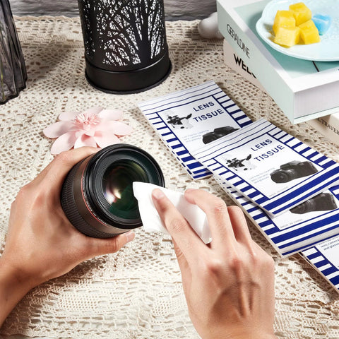 Lens cleaning tissue sheets and double-sided cloth kit; hands wipe a camera lens with tissue, 2 blue-striped booklets nearby.