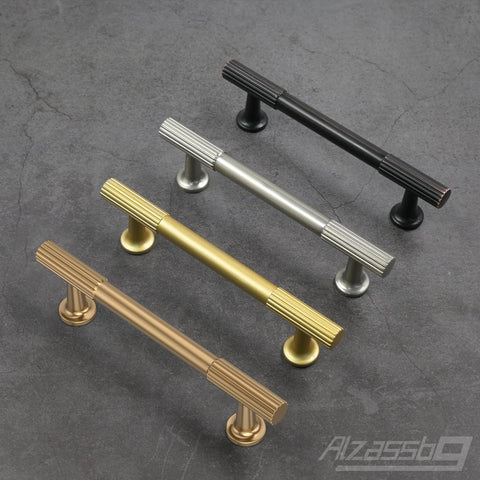 Four metal cabinet pulls in different finishes including champagne bronze, arranged on a gray surface, showcasing design and style.