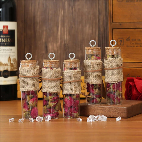Five glass vials filled with dried rose petals, wrapped in burlap, with cork tops, displayed on a wooden surface.