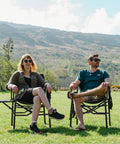 Two people sit on a Heavy Duty Oversized Camping Directors Chair With Side Table And Built In Pocket in a grassy field.
