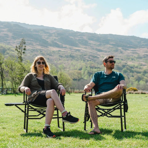 Two people sit on a Heavy Duty Oversized Camping Directors Chair With Side Table And Built In Pocket in a grassy field.