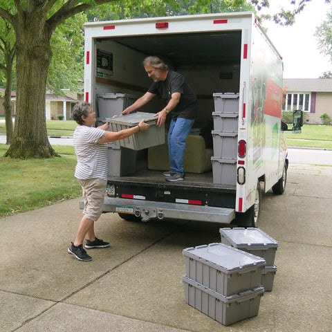 Two people loading Industrial Plastic Storage Tote With Hinged Attached Lid 2 Pack into a truck, driveway setting.