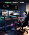 Person gaming at Dual Motor 3-Stage Standing Desk Frame With Electric Height Adjustment, Sit Stand DIY Desk Legs with monitors