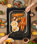 Top view of a table with assorted grilled seafood and steak, hand holding skewers over a grill surrounded by various dishes.