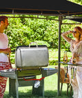 Outdoor scene with 5x8 Grill Gazebo With Extra Shade Awning Party Ready Vibe All Weather Steel Frame Double Tier Canopy Roof Sturdy Metal Serving Shelves Built In LED Lights Bottle Opener Patio Backyard BBQ Shelter Black, showing two people enjoying a barbecue.