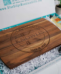 Personalized wooden cutting board with engraved names and date in circular design, brown finish, in gift box.