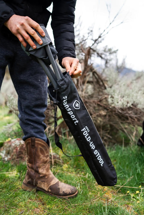 Person placing Apex Fold-Up Stool G-140 Black Grey Ultra Portable Camp Seat into carry bag outdoors