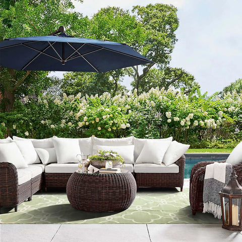 Outdoor seating area with white cushions and wicker furniture under a blue umbrella, set in a lush garden environment.