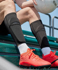 Soccer Shin Guard Sleeves With Insert Pocket And Top Lid, Breathable Mesh, Machine Washable, Keeps Shin Guards Locked In Place, 1 Pair, Black, Size Large Great For Training Sessions And Game Day Use worn by a player sitting with a soccer ball