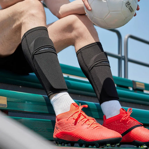 Soccer Shin Guard Sleeves With Insert Pocket And Top Lid, Breathable Mesh, Machine Washable, Keeps Shin Guards Locked In Place, 1 Pair, Black, Size Large Great For Training Sessions And Game Day Use worn by a player sitting with a soccer ball