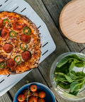 Freshly cooked pizza on 14 Inch Perforated Pizza Peel With Long Handle Aluminum Lightweight Pizza Paddle, surrounded by ingredients.