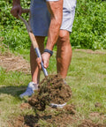 Person using Heavy Duty 43 Inch Round Shovel With Wooden D Handle And Stainless Steel Blade Built Tough For Digging Transplanting Planting Shrubs Snow Shoveling And Outdoor Yard Work in a garden.