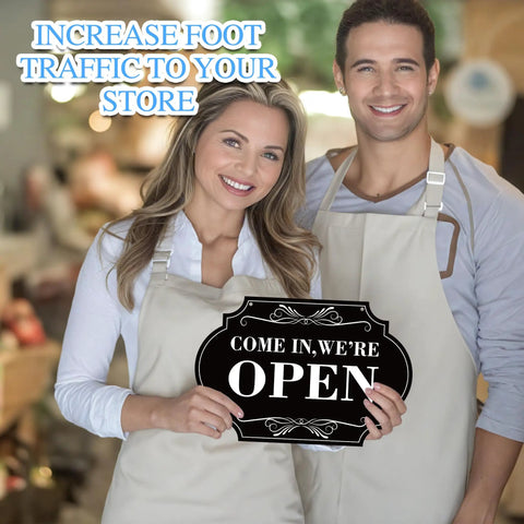 Two people in aprons holding a black open sign in a store setting.