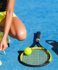 Person tying shoelaces next to a blue 32 oz insulated water bottle with straw spout and handle lid, tennis racket, and ball on court