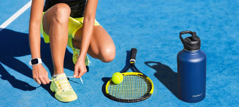 Person tying shoelaces next to a blue 32 oz insulated water bottle with straw spout and handle lid, tennis racket, and ball on court