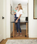 Woman holding baby standing beside Woodgrain Steel Baby Gate With Auto Close For Doorways, Adjustable From 29.75 To 40.5 Inches Wide And 30 Inches Tall, Brown Safety Gate That Looks Like Real Wood, Durable, Kid Friendly, Climb Resistant Barrier For Busy Homes.