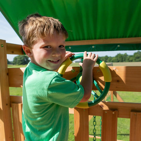 Child playing with steering wheel on Cedar Wood Swing Set With Wave Slide And Elevated Play Deck, Double Rock Wall, Rope Ladder.