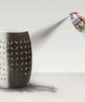 Hand spraying Badass Hammered Spray Paint In Sleek Dark Bronze on decorative metal vase with intricate cutouts.