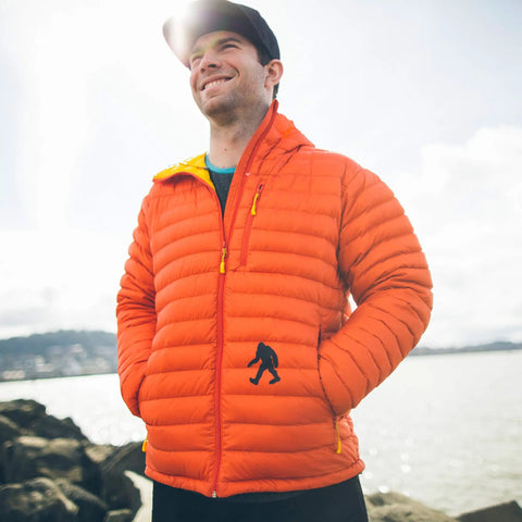 Person wearing an orange puffer jacket outdoors near a body of water with a Bigfoot silhouette on the pocket.