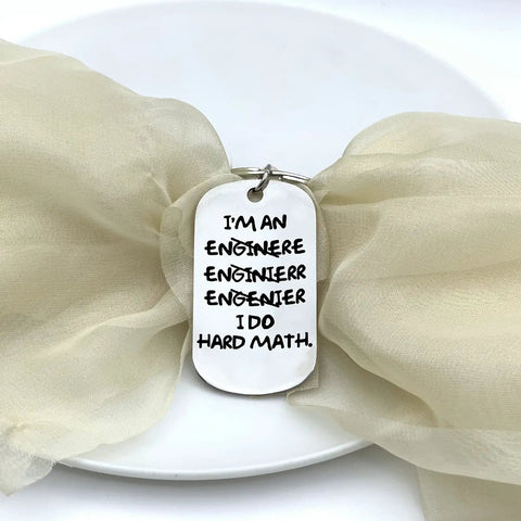 Engineering Keychain For Men A Funny Mechanical Gift For Engineers with engraved humorous text on a metal tag against fabric.