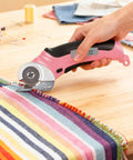 Heavy Duty Cordless Electric Scissors With Fast Usb Rechargeable Power And Self Sharpening Steel Blades For Slick Cuts Through Denim Fabric Leather Carpet And Stubborn Cardboard In Nifty Pink cutting striped fabric on a wooden table.