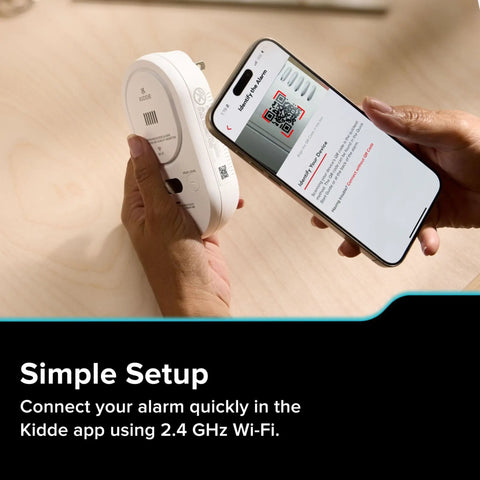 Person setting up a Smart Carbon Monoxide Detector And Indoor Air Quality Monitor With Real Time Alerts No Subscription Needed Plug In With Battery Backup 2 4 GHz Wi Fi Rotating Plug 85 dB Alarm IAQ Insights For Homes using a smartphone with the Kidde app on a wooden table.