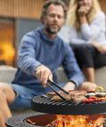 Man grilling food on a 35 Inch Outdoor Fire Pit Table With 360 Degree Rotating Grill, Wood Burning Fire Pit With Lid And Poker