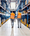 Two workers in orange safety vests walking through a warehouse aisle filled with racks of boxes and products.