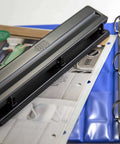 Black compact three-hole punch next to binder on desk, 8-sheet capacity, removable chip tray, ideal for office or home use.