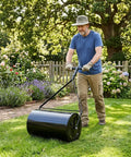 Man using Lawn Roller With Push And Tow Versatility, 14 By 24 Inch Roller, 16 Gallon Capacity, on lawn beside garden.