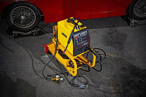 HEAVY DUTY YELLOW POWDER COATED STEEL WELDING CART WITH SMOOTH ROLLING CASTERS AND SLANTED WORK SURFACE BUILT TO CRUSH THE GRIND AND HAUL YOUR GEAR FAST positioned beside a red car.