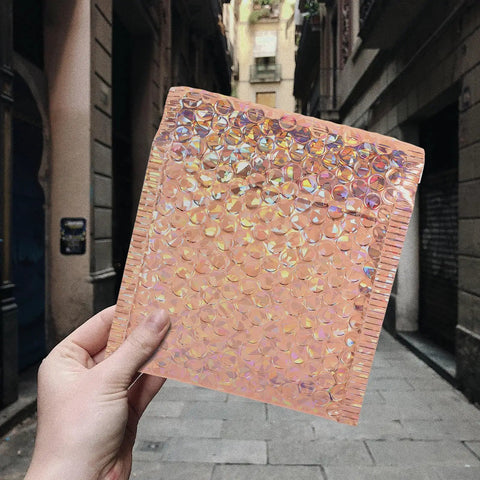 Hand holding a rose gold bubble mailer, part of Rose Metallic Padded Mailers 20 Pack 6x5 Inch Self Sealing Cushioned Shipping Bags For Mailing Clothes And Small Packages Durable Protective Packaging That Just Works For Quick Ship, in an urban background