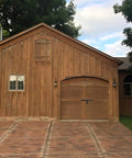 Wooden barn with a brick driveway, featuring an arched wooden gate door, surrounded by trees on a sunny day.