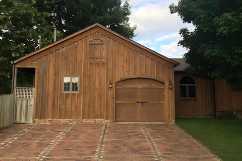 Wooden barn with a brick driveway, featuring an arched wooden gate door, surrounded by trees on a sunny day.