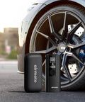 Portable Cordless Tire Inflator With Hard Case Bundle Quick Accurate Air Pump For Car Truck SUV Motorcycle And Bicycle 120 PSI Gauge Compact Air Compressor With Carrying Case next to car wheel.