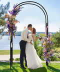 Bride and groom kissing under 7.2 Ft Outdoor Steel Garden Arch Arbor With Stakes, Wide Sturdy Black Metal Archway with floral decor.