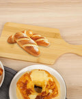 Bamboo cutting board with handle on a wooden table, 11x5 inches, for bread, cheese, and charcuterie.