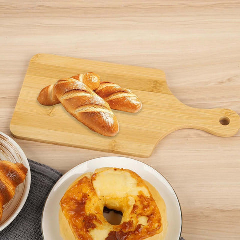 Bamboo cutting board with handle on a wooden table, 11x5 inches, for bread, cheese, and charcuterie.