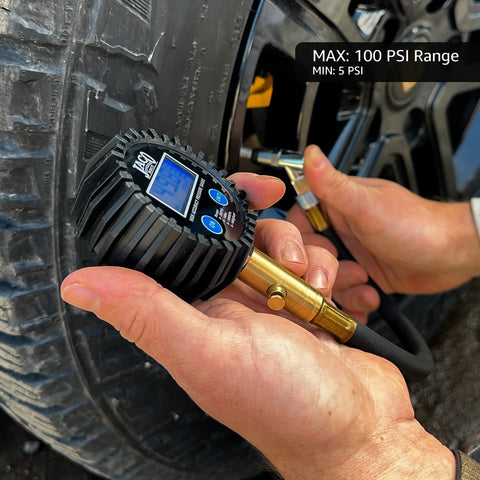Close-up view of JACO ElitePro digital tire pressure gauge with brass nozzle on a car tire; black ribbed body, blue LCD.