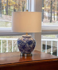 Blue And White Floral Ceramic Table Lamp With White Linen Shade And Brushed Nickel Accents 3 Way Lighting Ginger Jar Inspired Cozy Decor For Living Room Or Bedroom Soft Warm Glow And Timeless Charm on a wooden table by window.
